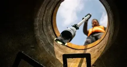 Person in a manhole, inspecting with a long camera tool. Sky visible.