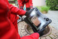 A man in a red jacket is holding a tablet in front of a manhole cover.