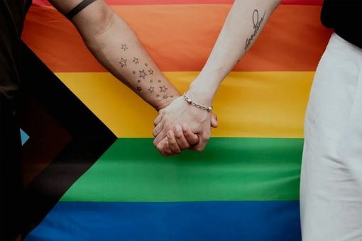 Two people are holding hands in front of a rainbow flag.