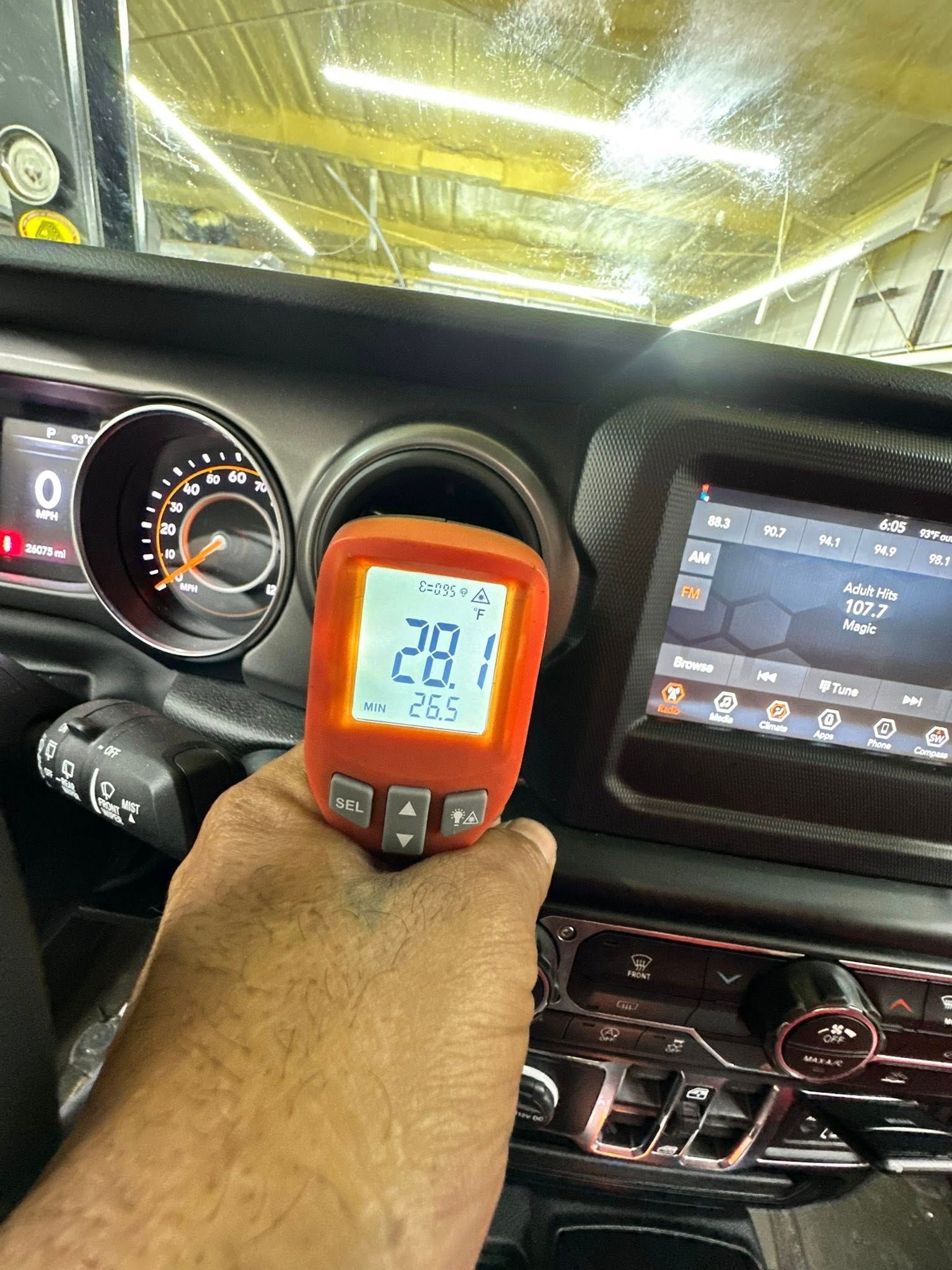 Hand holding an orange temperature gauge in a car vent, displaying 28.1 degrees, near dashboard displays.