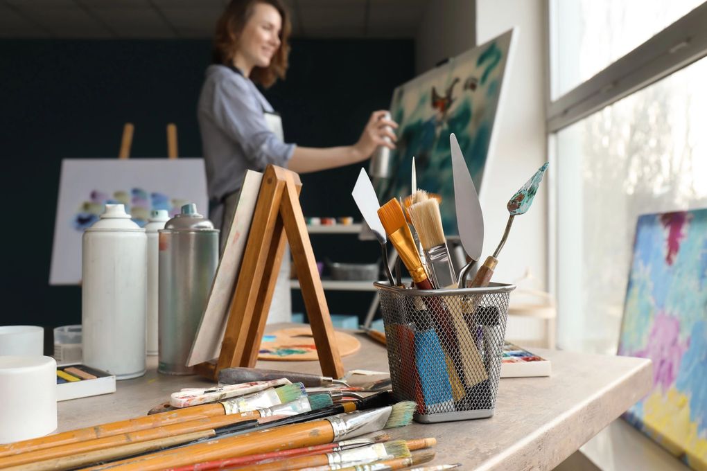Un ambiente da studio con pennelli e spatole in una tazza, e un artista che lavora su una tela sullo sfondo.