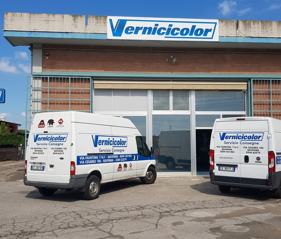 Due furgoni bianchi della Vernicicolor parcheggiati davanti a un edificio in mattoni con un'insegna aziendale dello stesso colore sulla facciata.