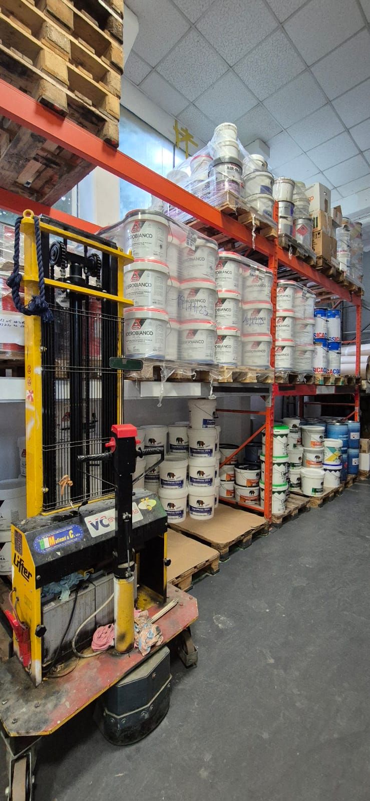 Un carrello elevatore giallo si trova in un corridoio di un magazzino, accanto a scaffalature industriali alte e piene di secchi di plastica bianca.