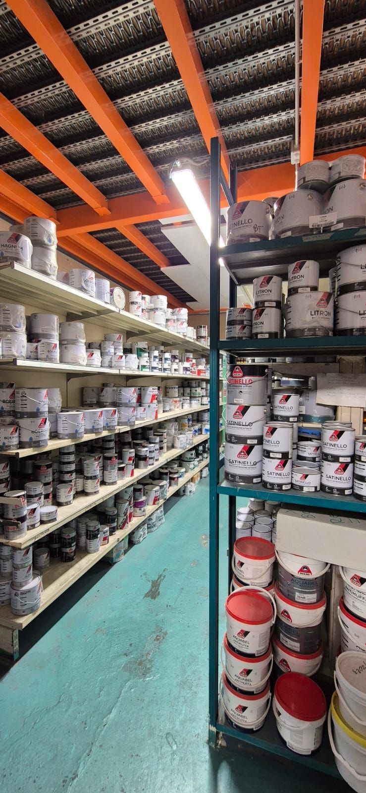 Uno stretto corridoio di un magazzino con alte scaffalature metalliche piene di lattine e contenitori di vernice di vario tipo.
