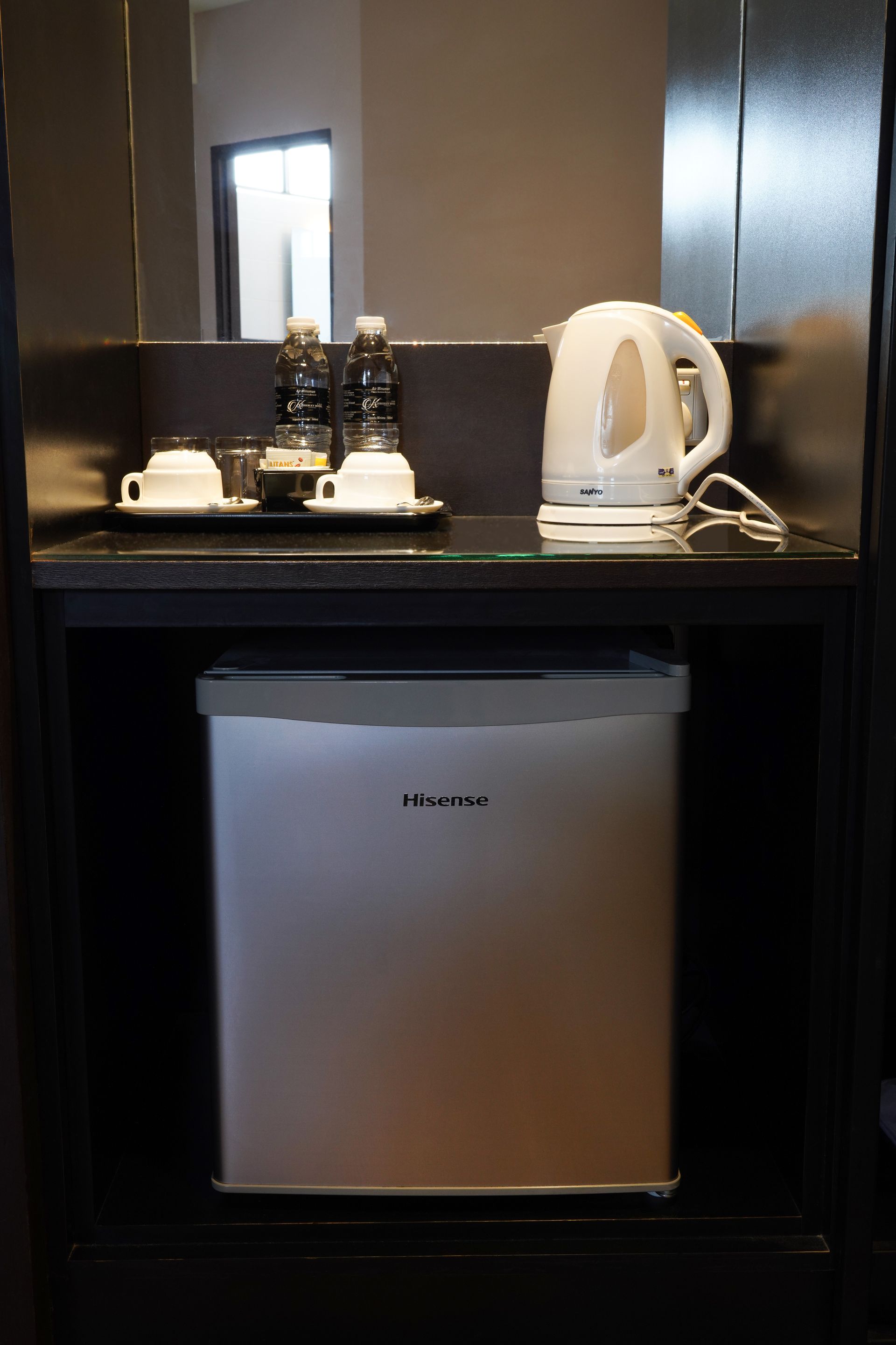A hotel room with a fridge , kettle , cups and bottles.