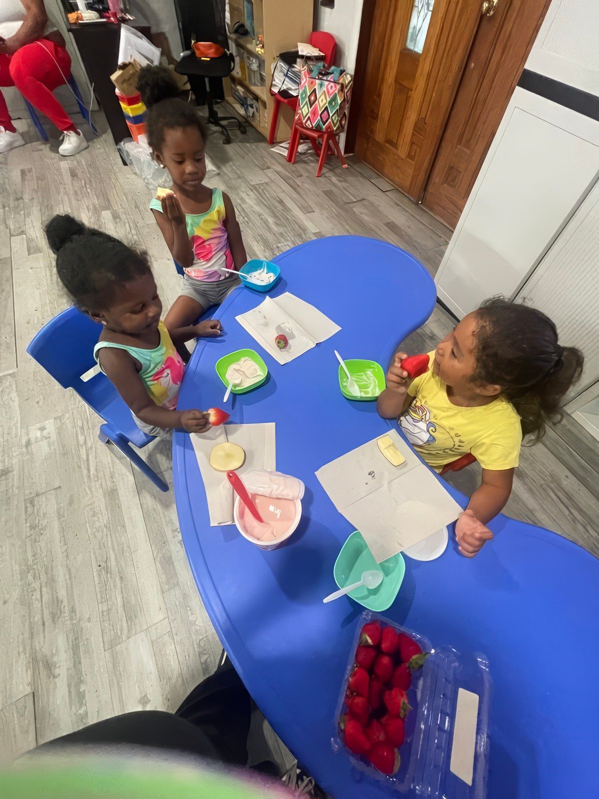 Children at blue table with fruit and snacks, in a brightly lit room.