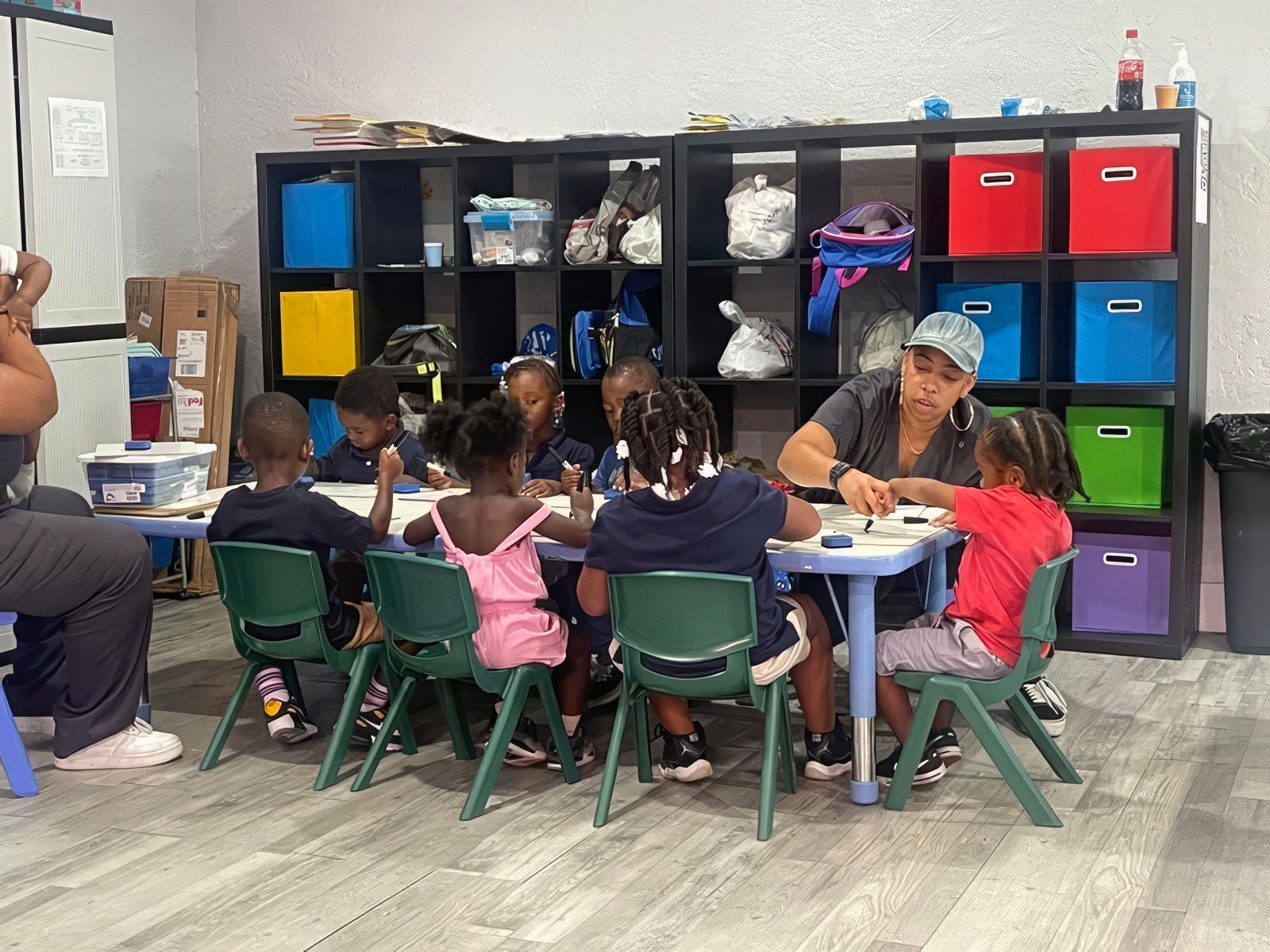 Children and a teacher at a table. They are sitting in green chairs. Shelves with colorful boxes are in the background.