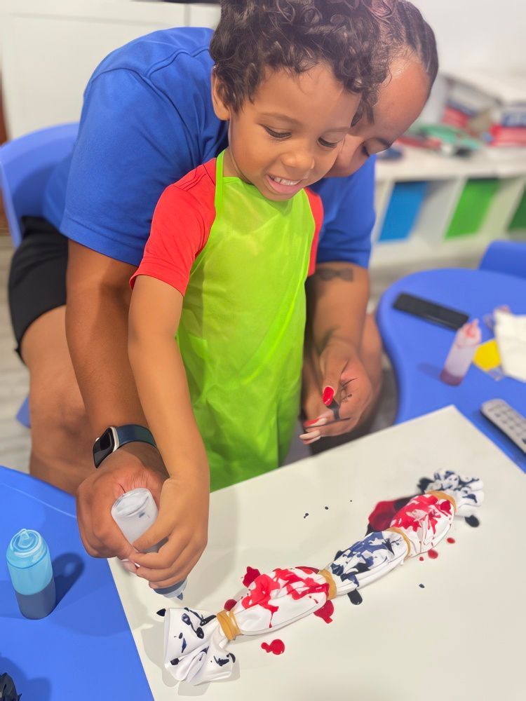Person and child tie-dyeing on a table, child wearing a smock, using a dye bottle.
