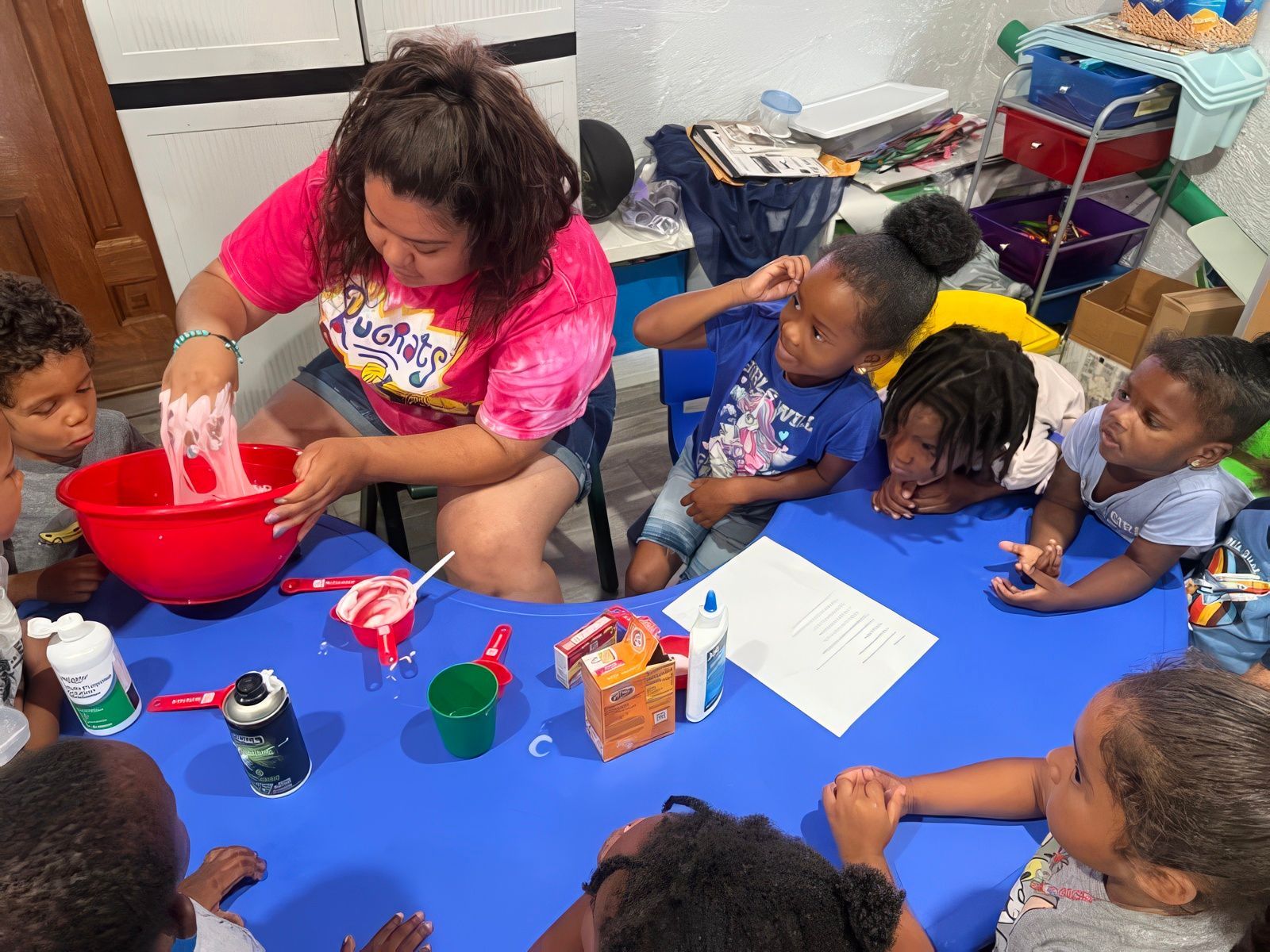 Woman making slime with children at a blue table; red bowl, bottles, and ingredients are present.
