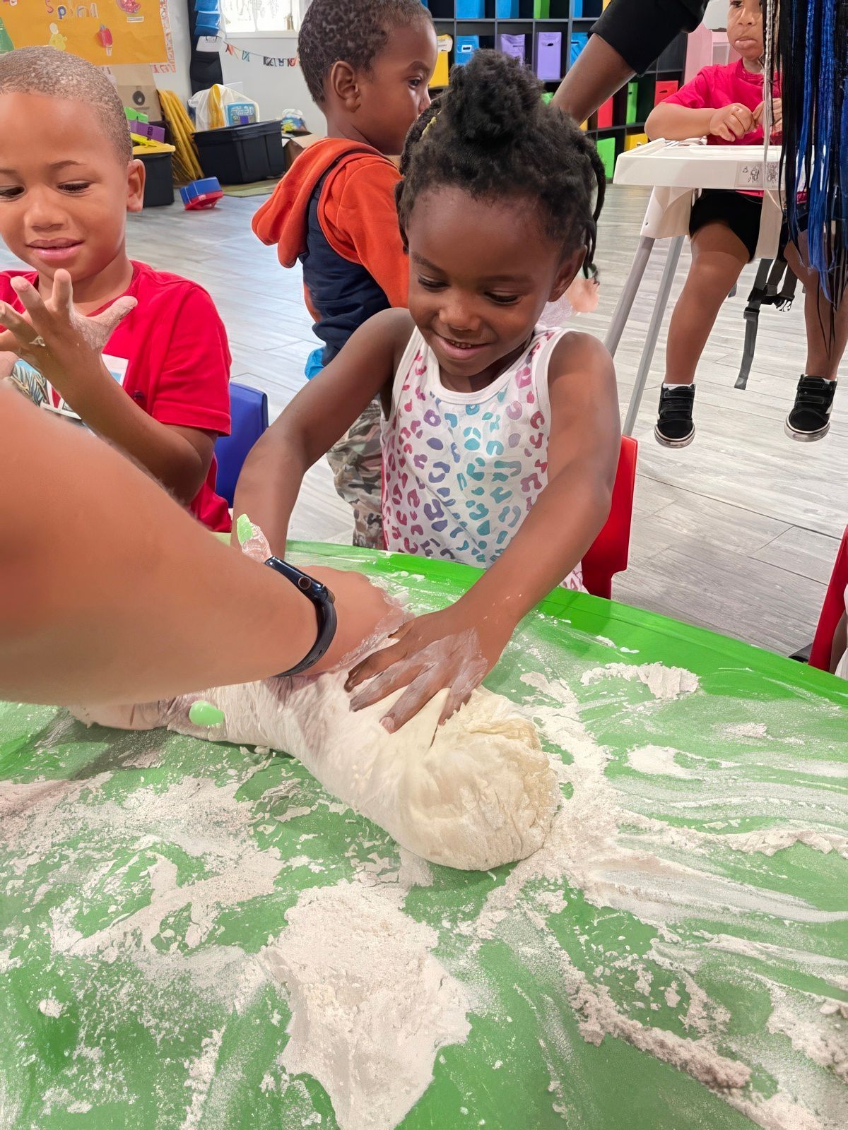 Children at a green table, hands-on with dough. Smiling child presses dough with a teacher. Flour is scattered.