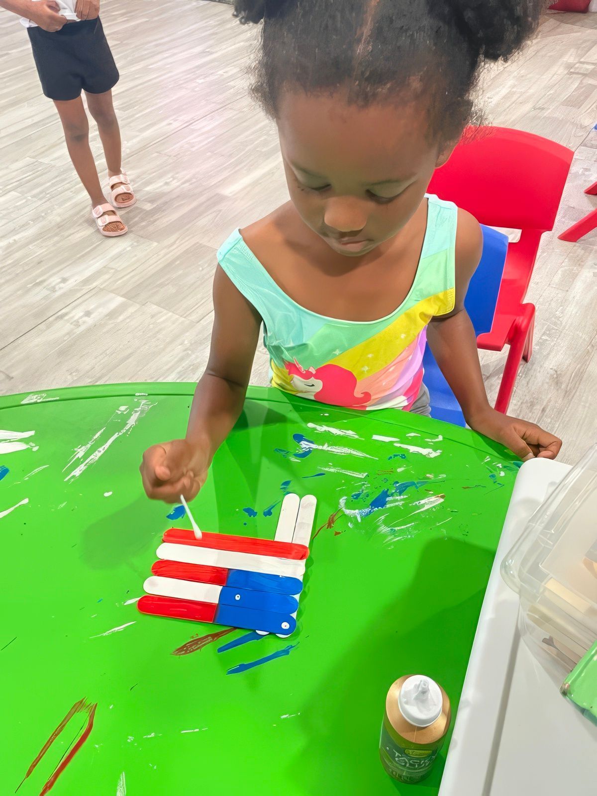 Girl painting a popsicle stick American flag on a green table.