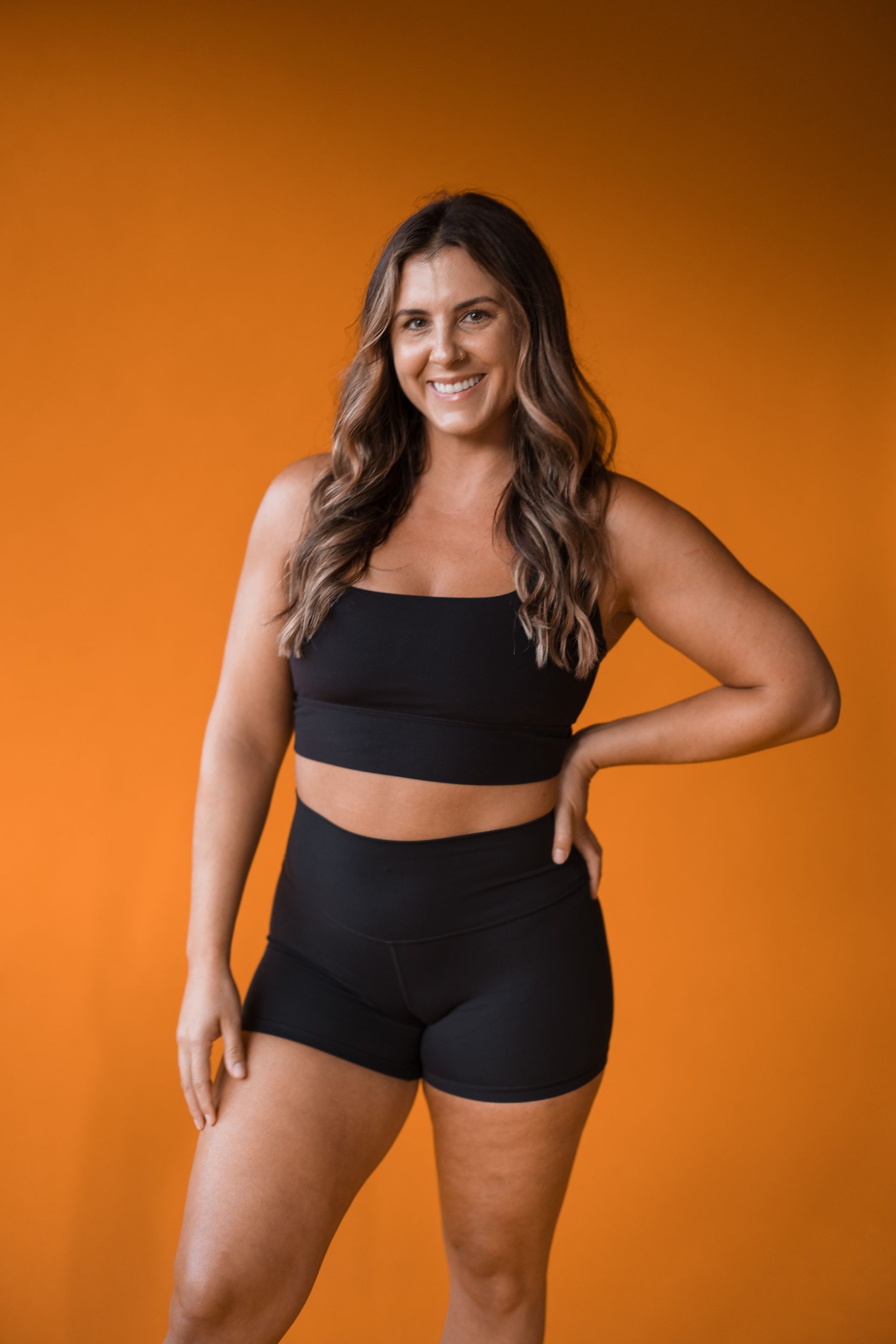 A woman in a black crop top and shorts is standing in front of an orange background.