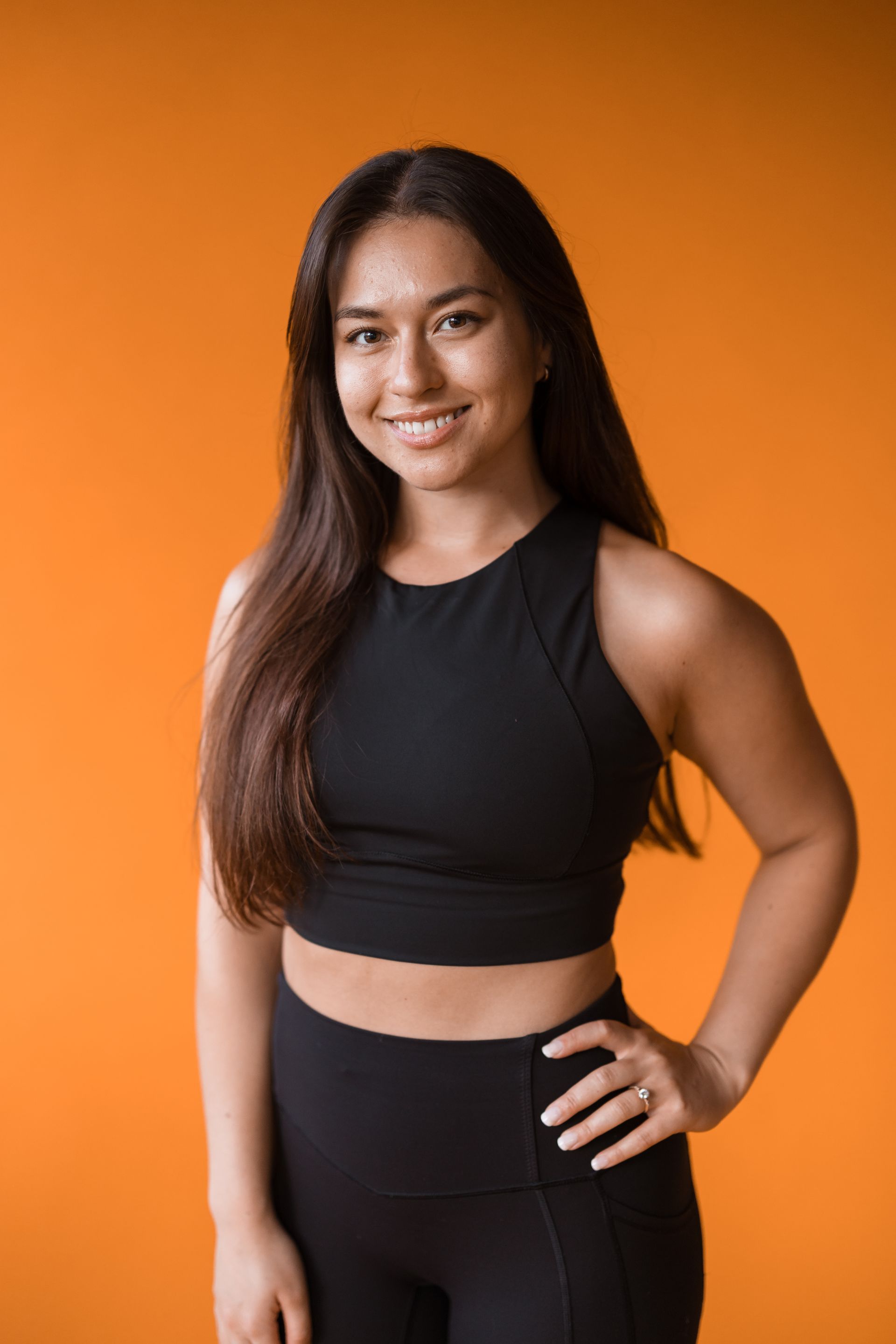 A woman in a black crop top and black leggings is standing in front of an orange background.