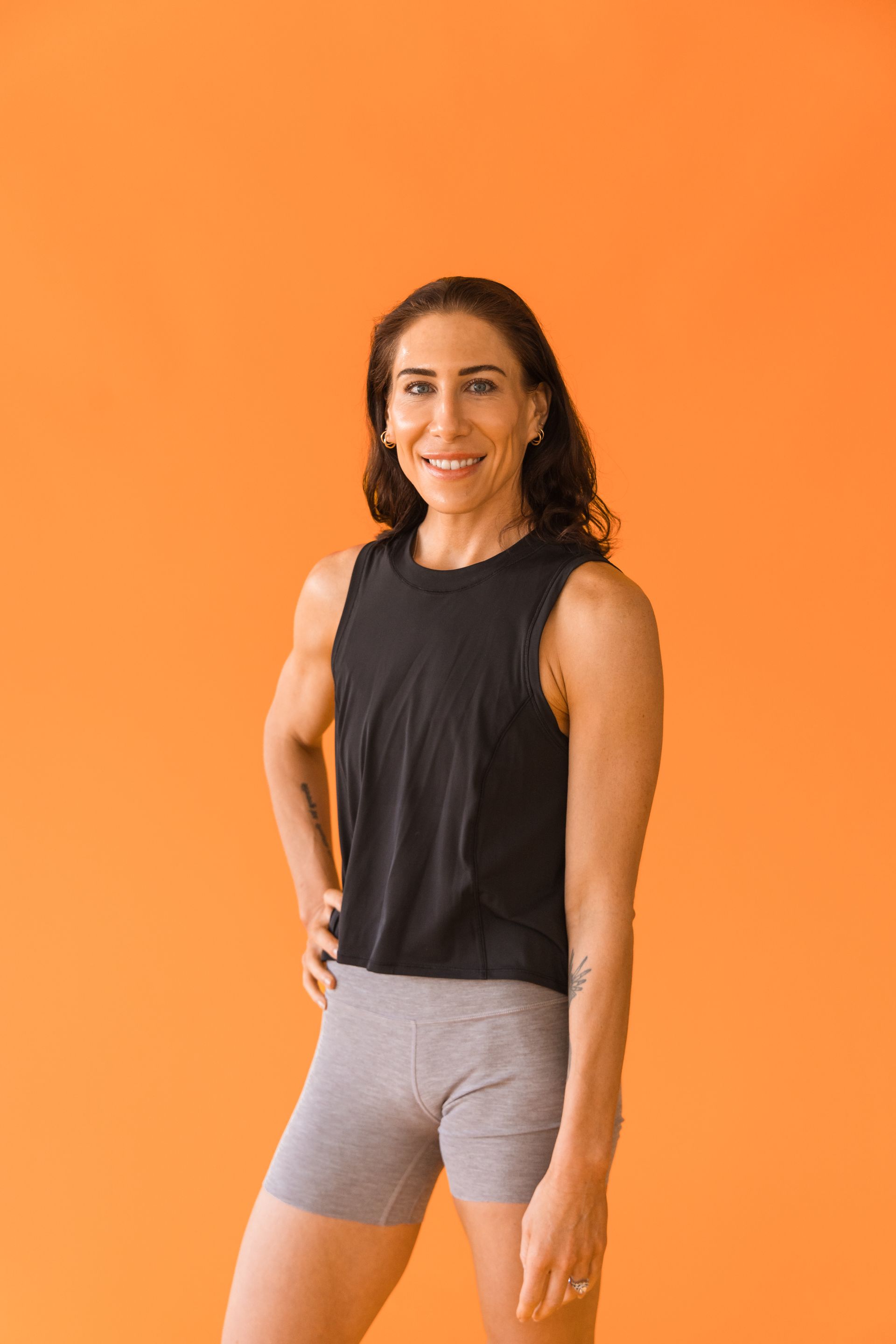A woman in a black tank top and grey shorts is standing in front of an orange wall.