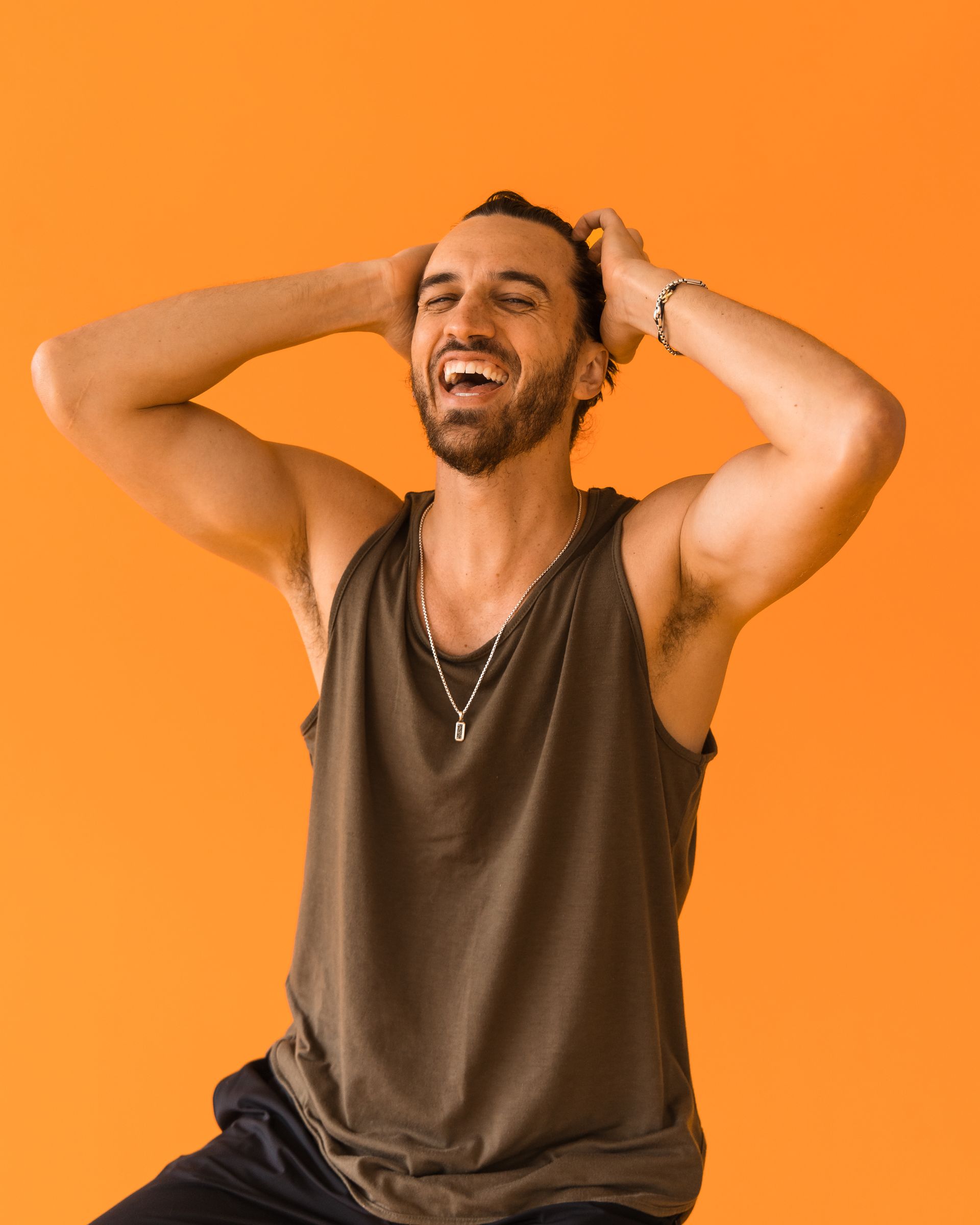 A man in a tank top is laughing with his hands behind his head.