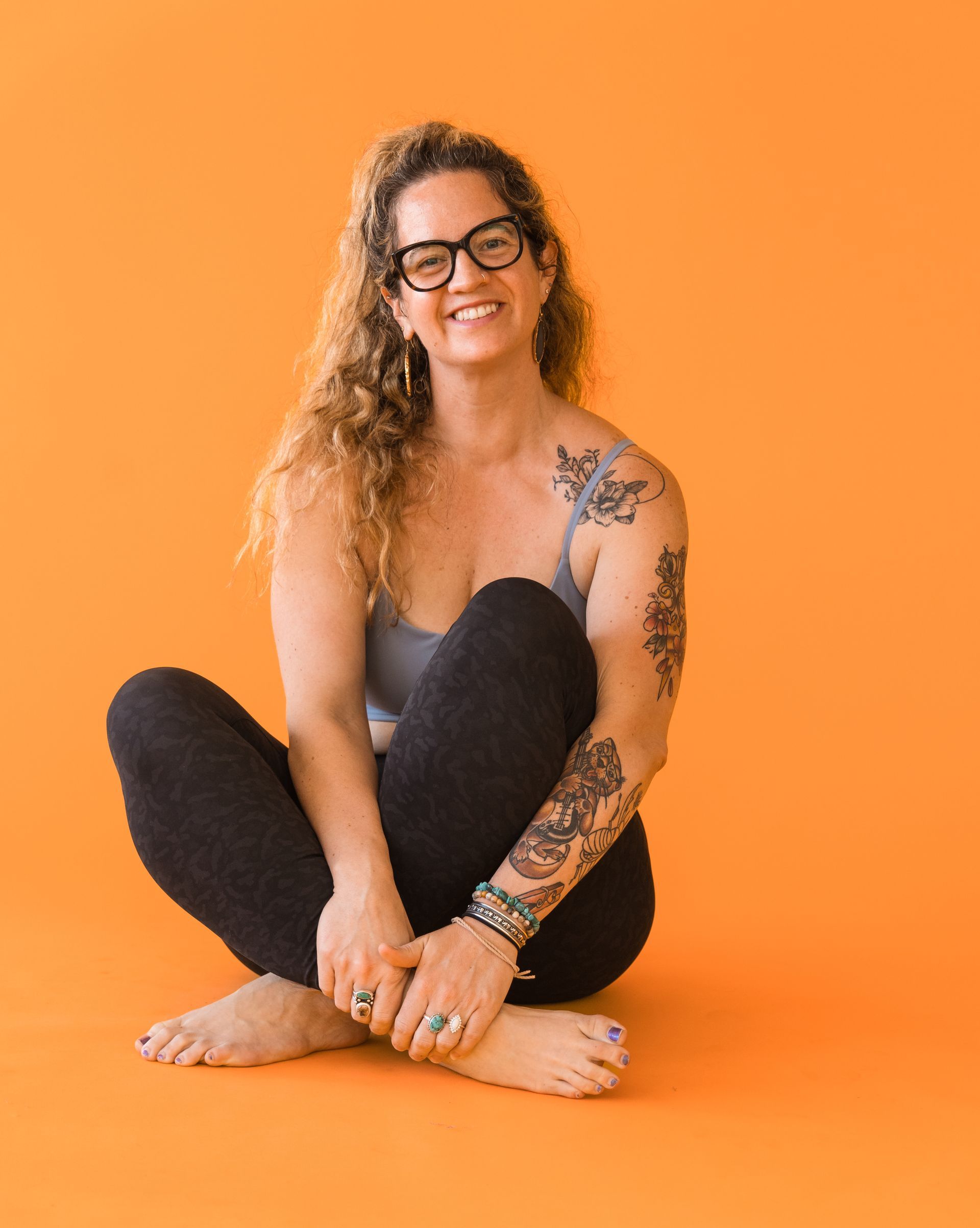 A woman with glasses and tattoos is sitting on the floor with her legs crossed.