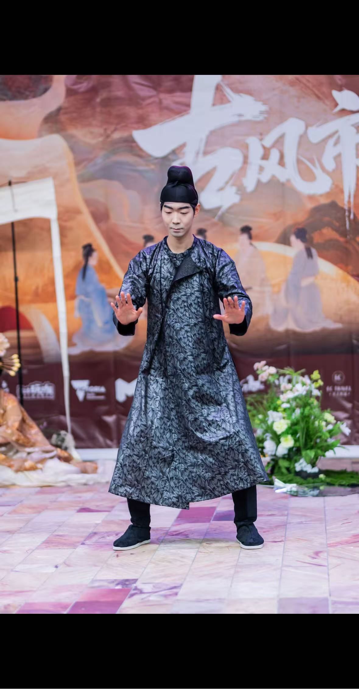 Man in black, traditional attire, dancing. Stage background with floral decorations.