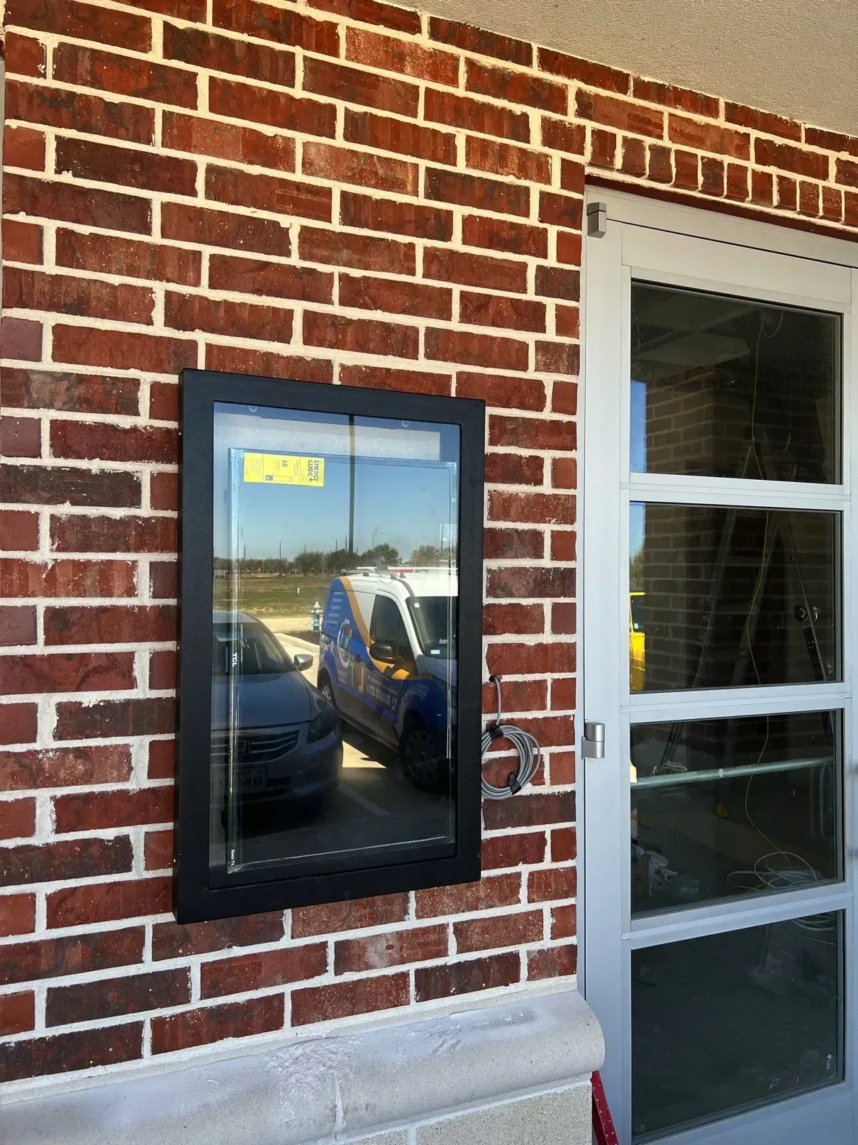 Brick building exterior with a drive-up window and a clear glass door. A car and a van are reflected in the window's glass.