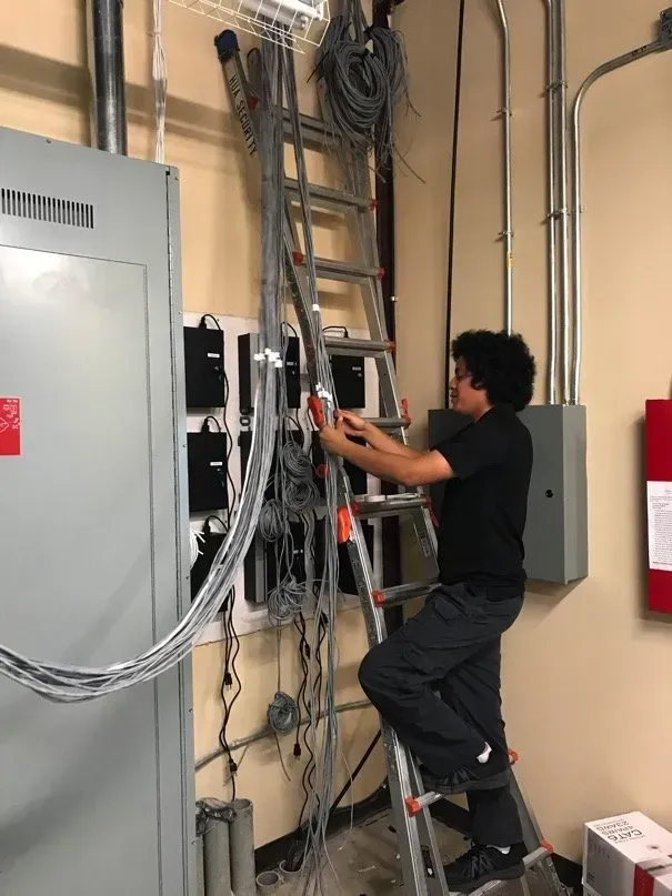 A person on a ladder works on a tangle of wires in an electrical room. They are wearing a black shirt and gray pants.