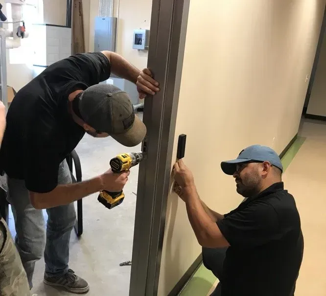 Two men in a room installing a door frame. One uses a drill while the other holds a piece.