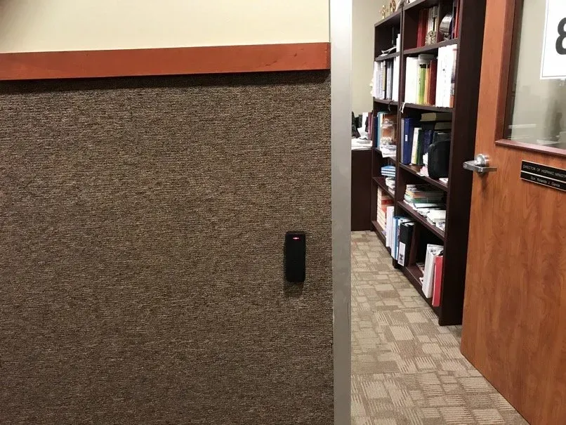 A hallway with a brown carpeted wall and a door to an office with bookshelves. A black access control reader is mounted on the wall.