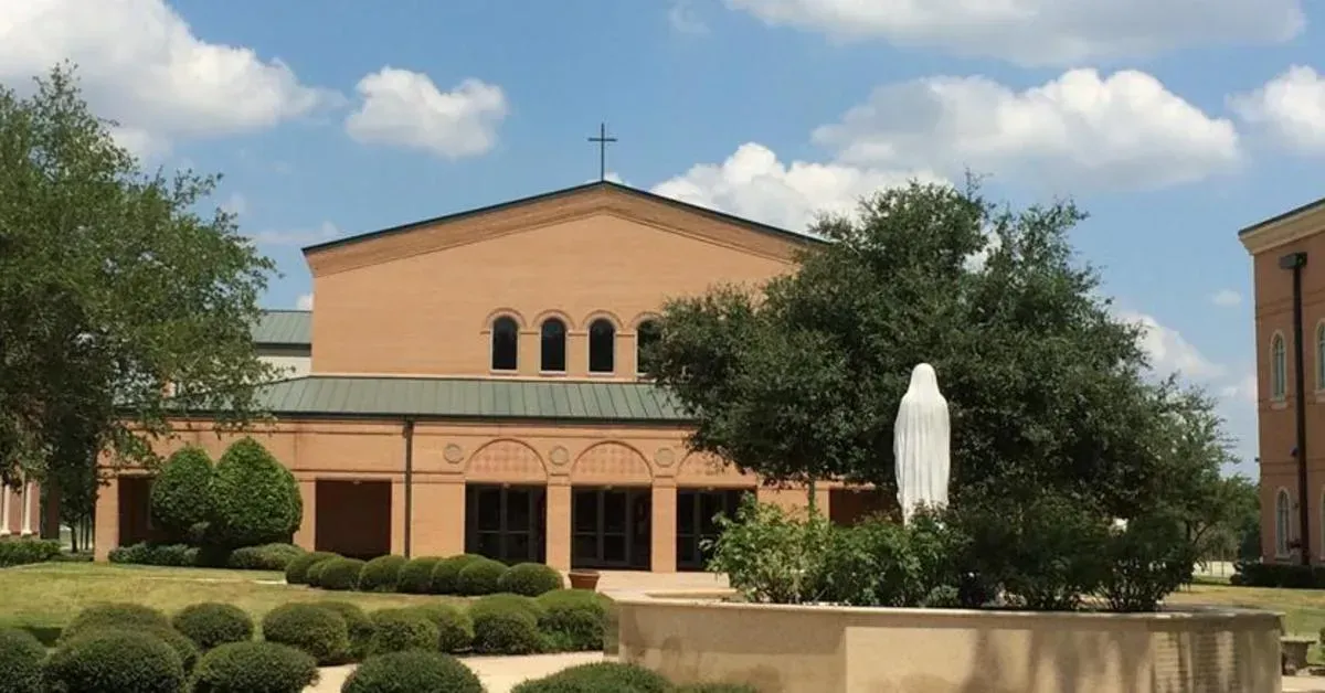 A church building with a cross on top, a statue of Mary, and a blue sky with clouds in the background.