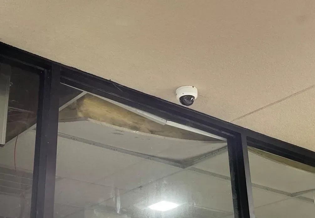 Security camera mounted on a ceiling above large glass windows with black frames. A damaged ceiling section is visible.