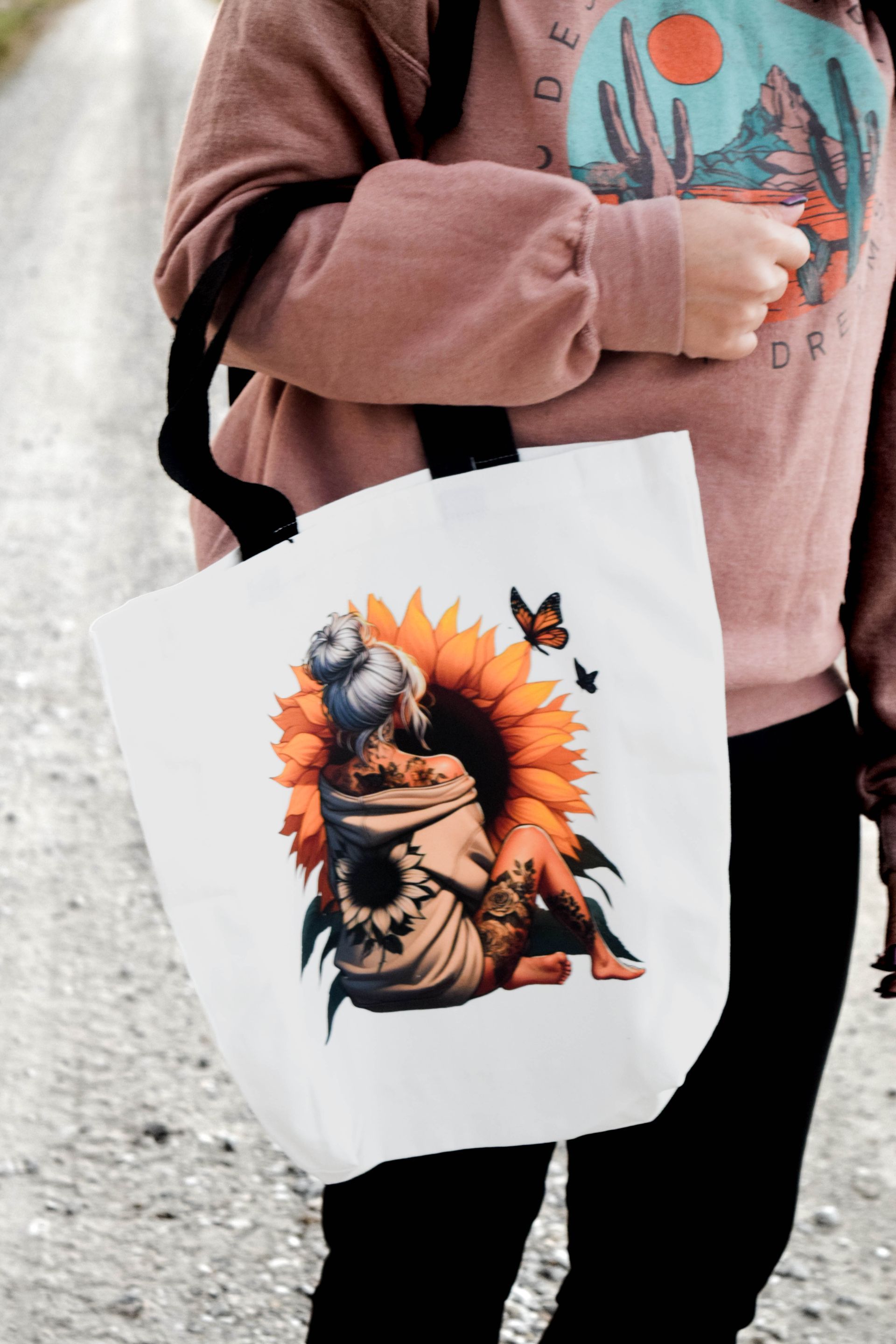 Woman holding a white tote bag with a sunflower design. She is wearing a pink sweater and black pants.