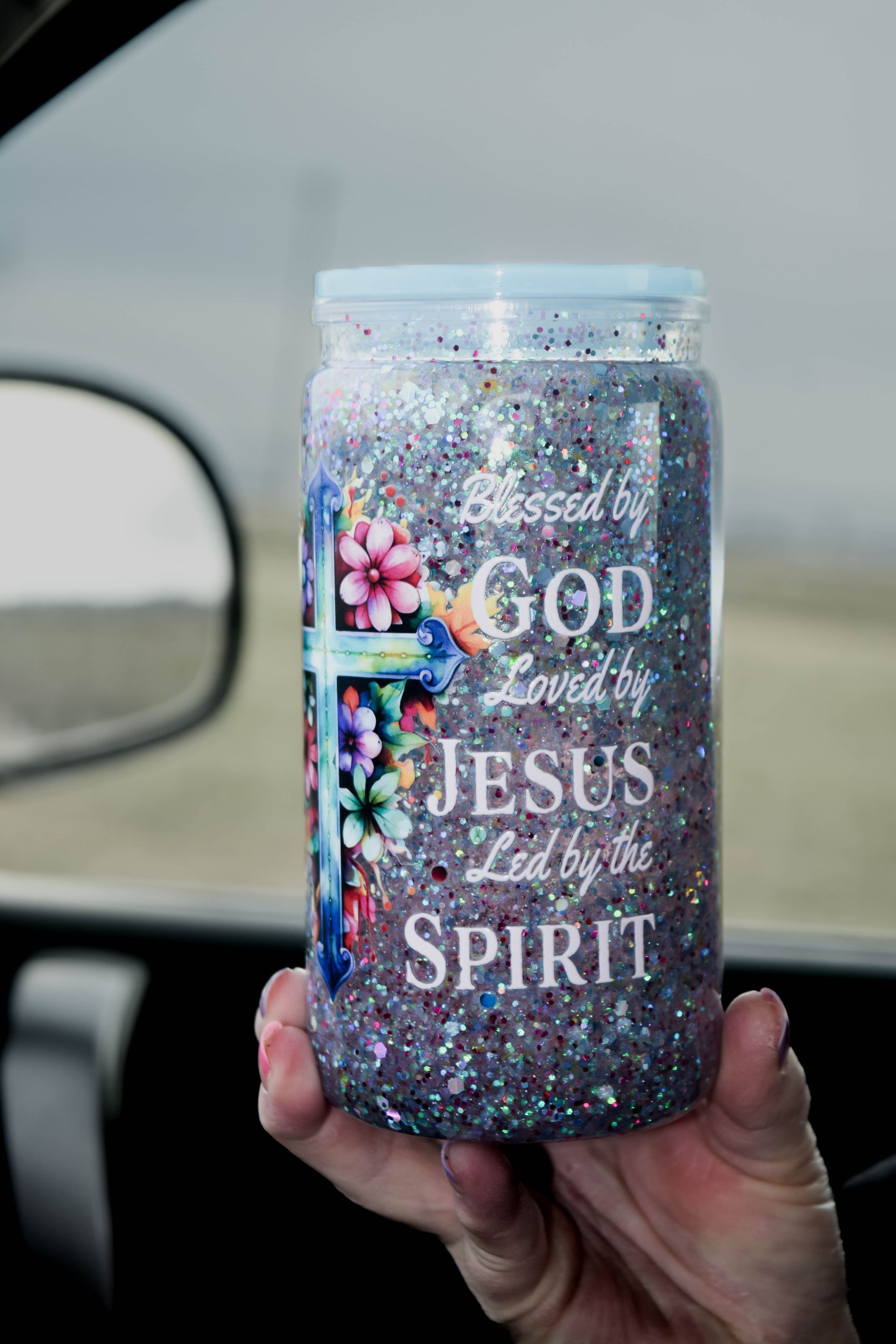Hand holding a glitter-filled tumbler with a cross and floral design, text: 