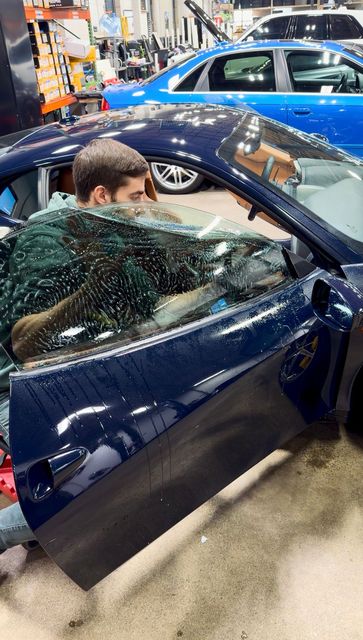 Man sitting in a dark blue car at an auto show, with the driver’s door open and other cars in the background