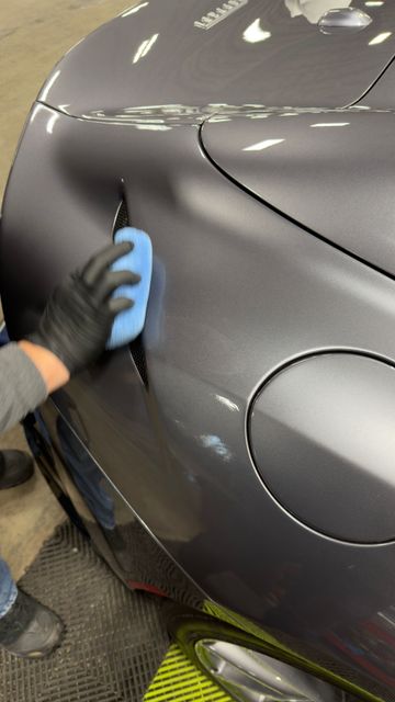 Gloved hand wipes the dark gray car’s rear quarter panel with a blue cloth in a garage.