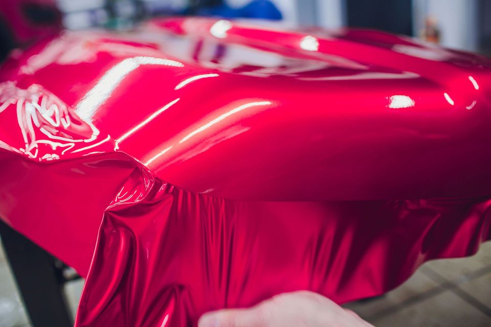 Glossy red car being wrapped in vinyl film, with hands smoothing the material across the body panel