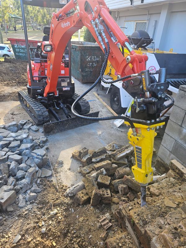 Orange excavator with a jackhammer breaking bricks in an outdoor construction setting.
