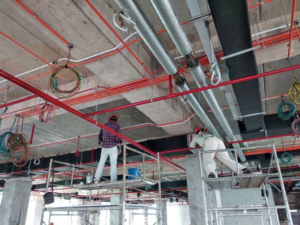 Dos trabajadores en un andamio instalando tuberías y cableado en el techo de una obra en construcción.