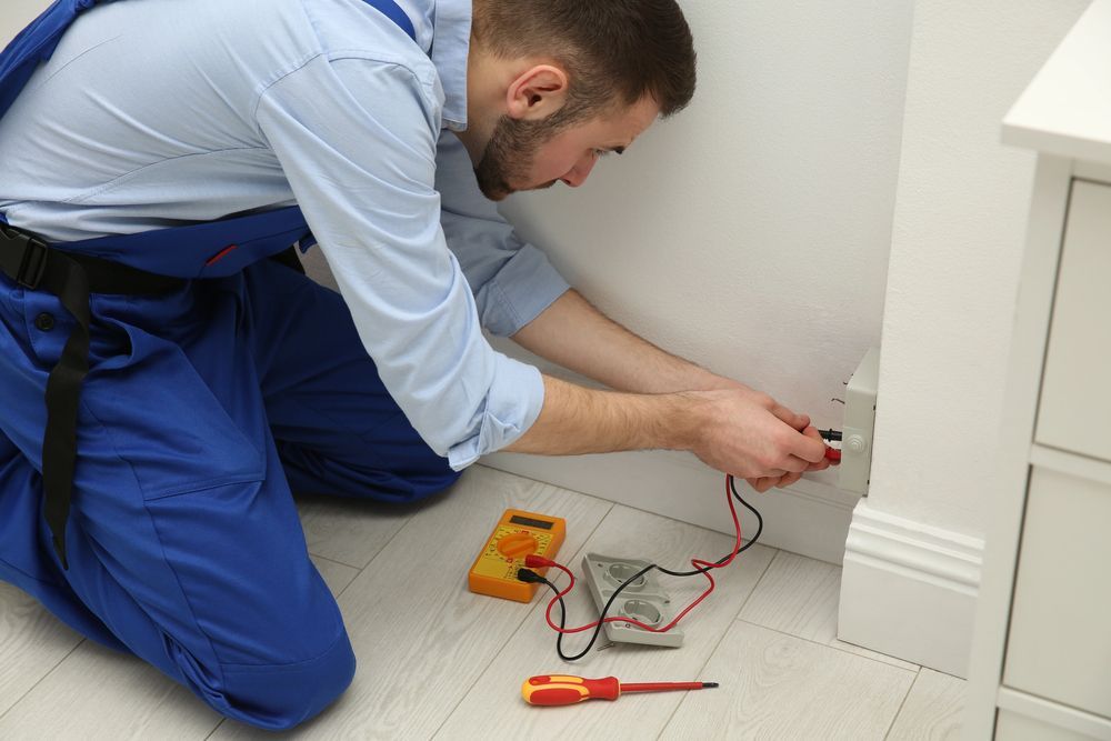 Un electricista con mono azul prueba un tomacorriente con un multímetro en una habitación blanca.