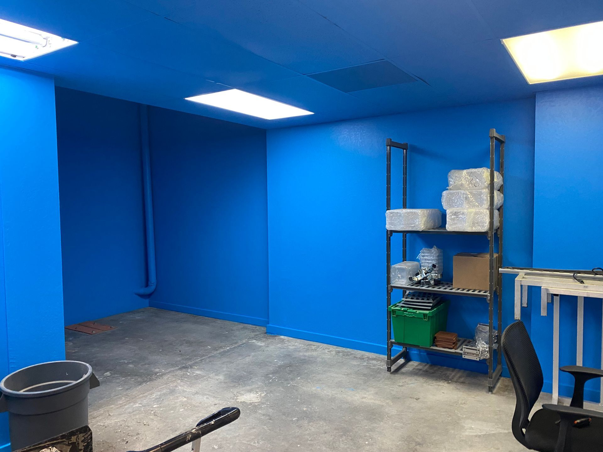 Blue empty room with concrete floor, shelves, buckets, and a chair under bright ceiling lights
