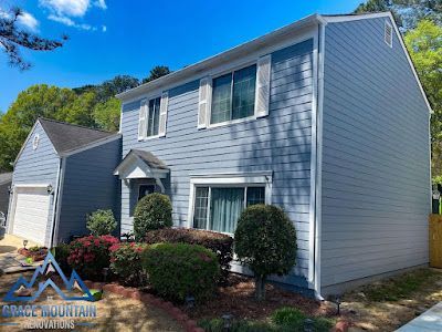A house with a blue siding and white trim is sitting on top of a lush green hillside.