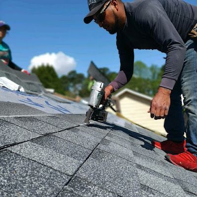 Man in sunglasses using a nail gun to install shingles on a roof. Another person is nearby on the roof.