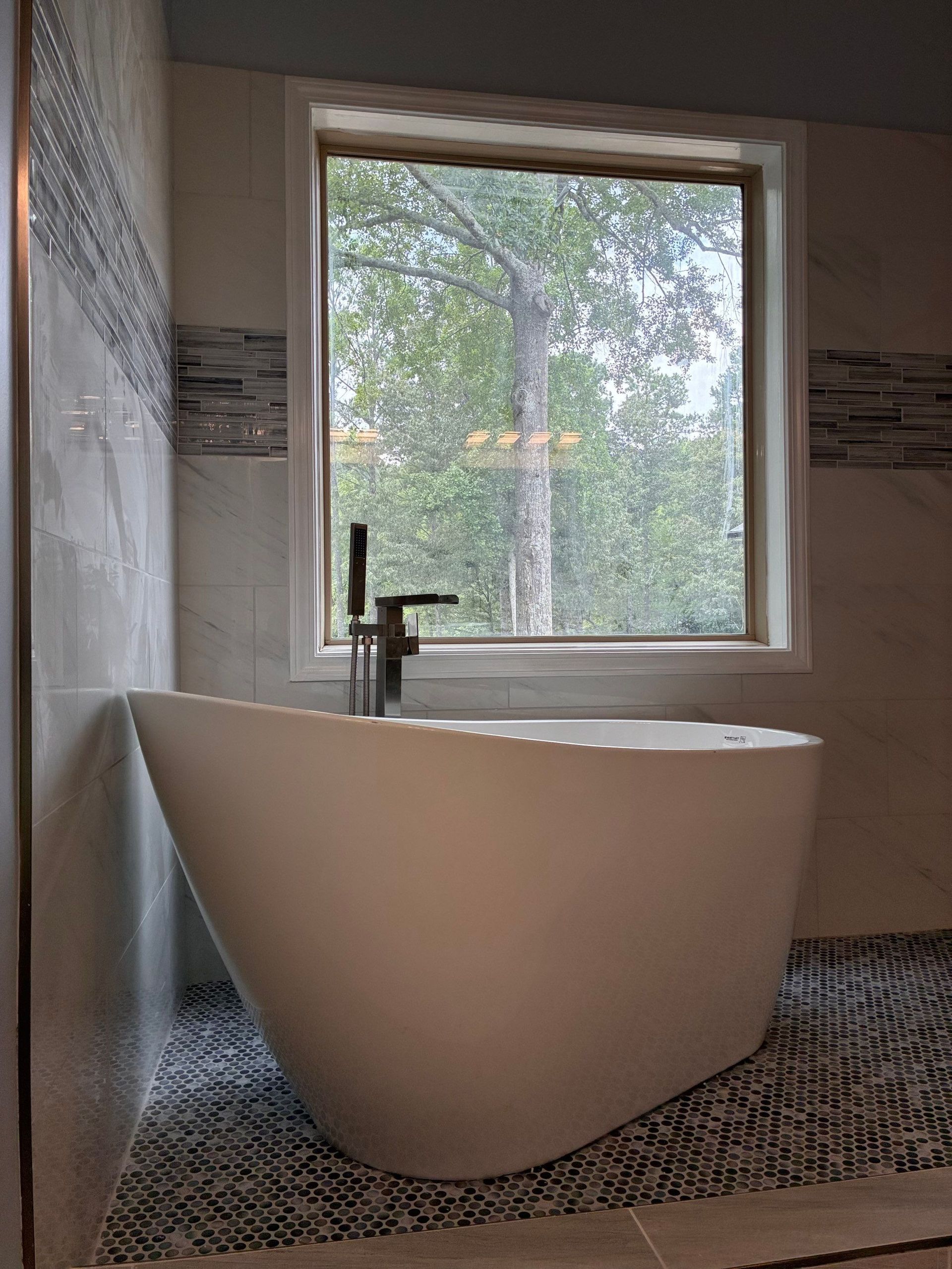 Modern white bathtub near a window overlooking trees, set in a bathroom with mosaic tile flooring.