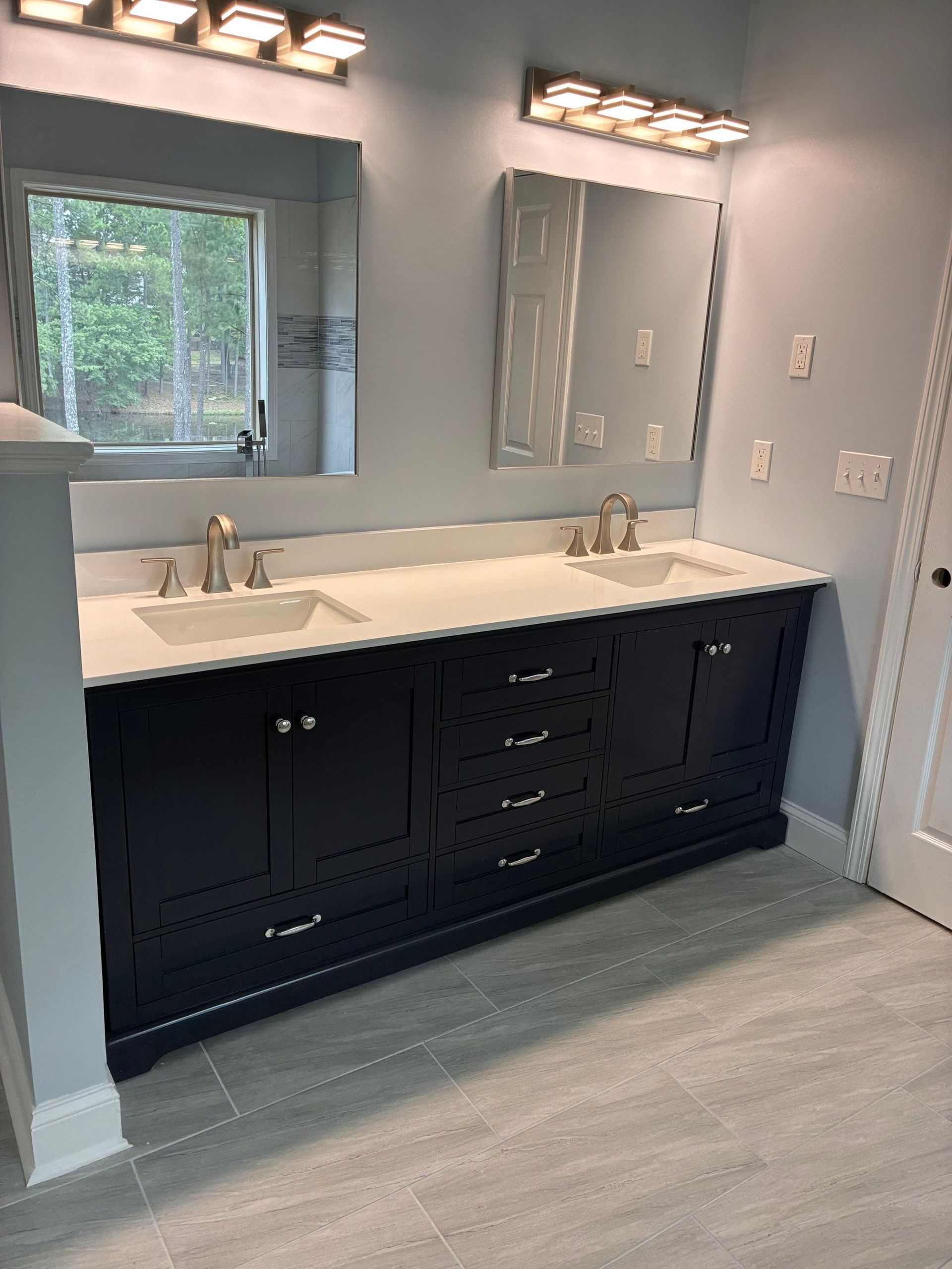 Bathroom with dark blue vanity, white countertop, two sinks, mirrors, light fixtures, and light blue walls.