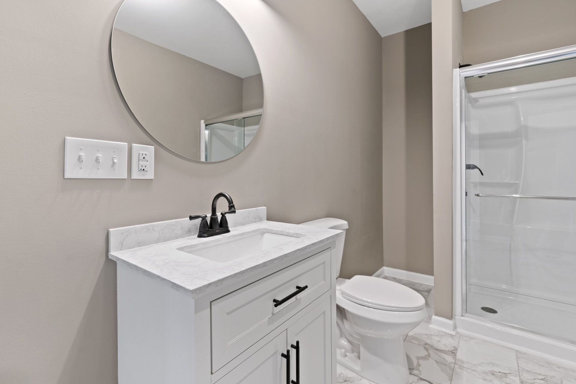 Bathroom with white vanity, toilet, and shower. Round mirror, black faucet, light beige walls.