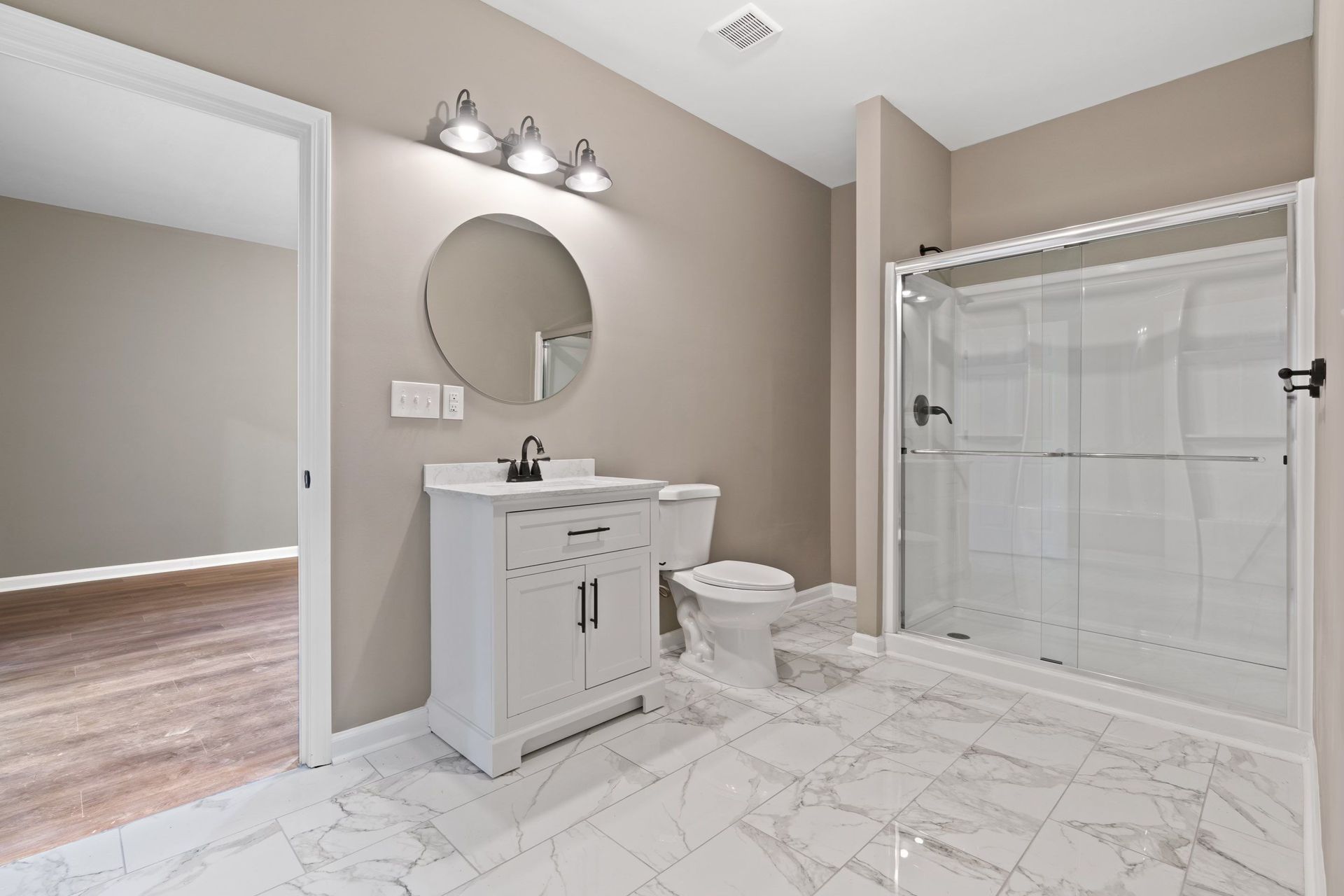 Bathroom with white vanity, toilet, and shower. Beige walls, round mirror, and marble tile.