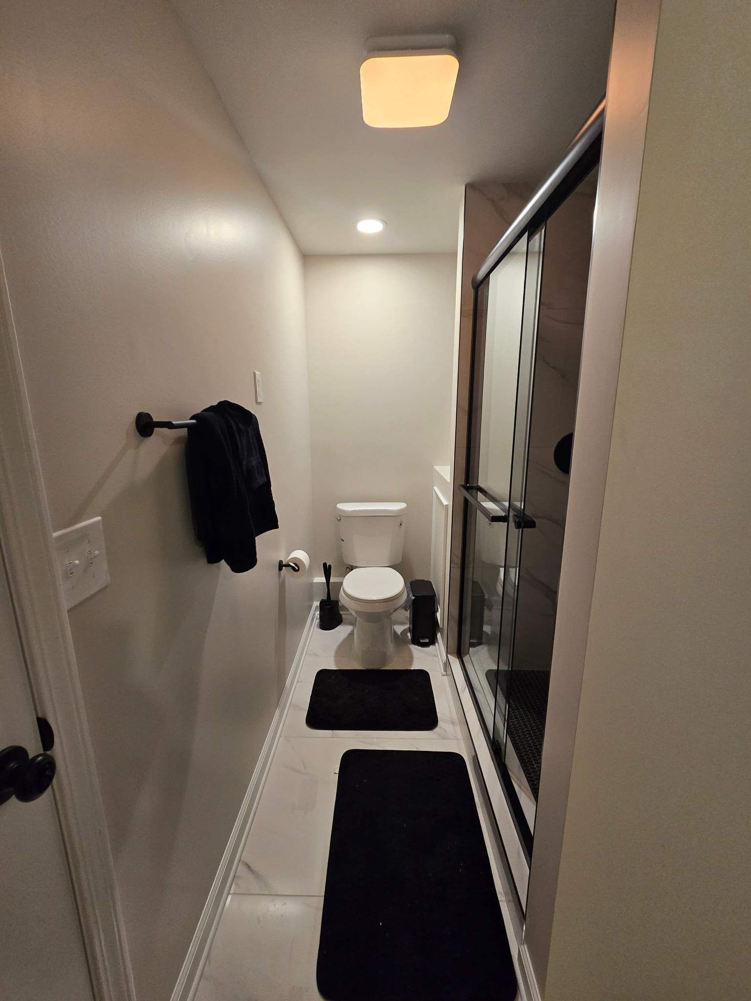 Narrow bathroom with toilet, shower, and black accents. Light-colored walls and flooring.