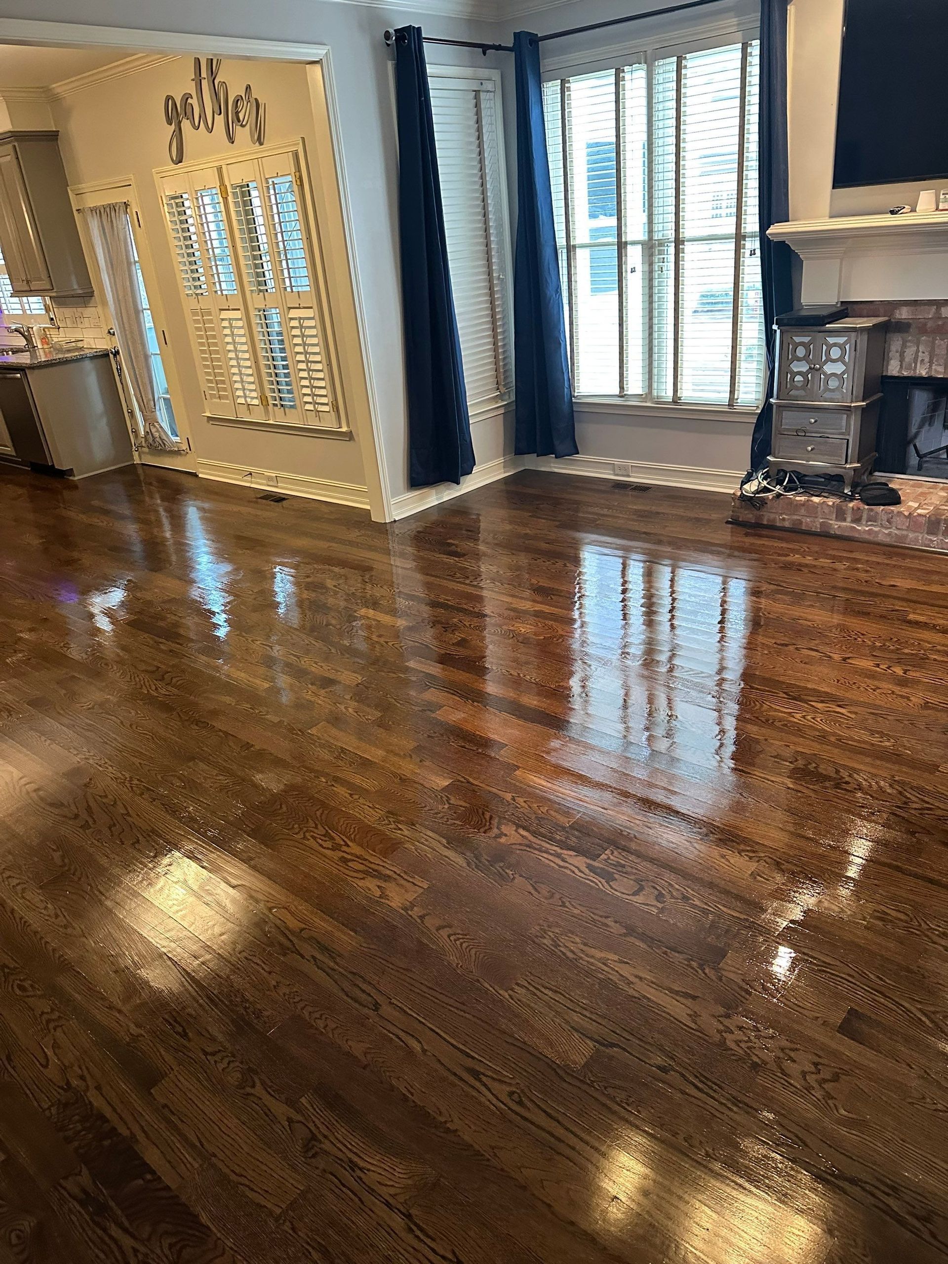 Shiny, dark hardwood floors reflect light in a living room with a fireplace, window, and blue curtains.