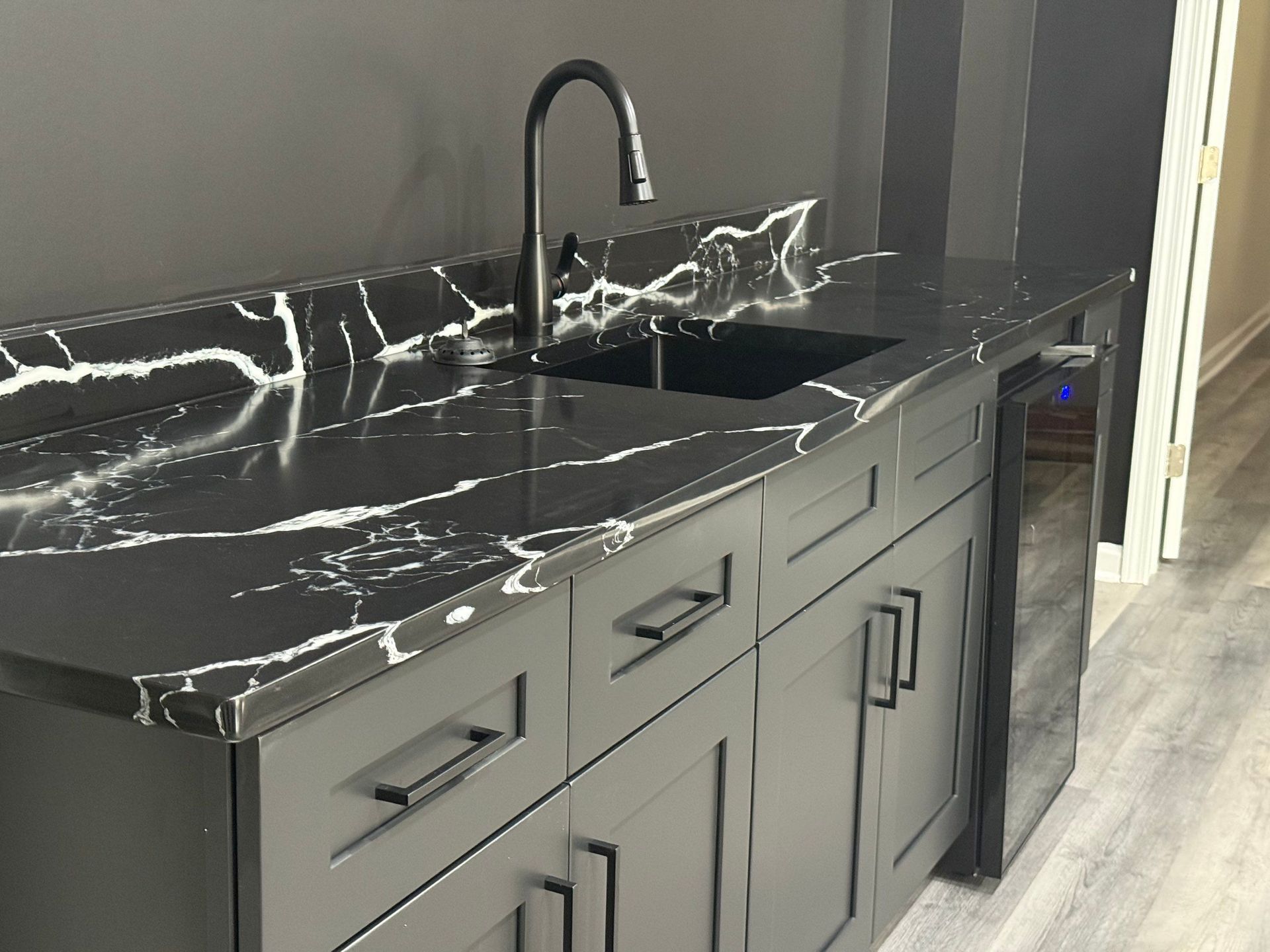 Dark gray wet bar with black and white countertop, black faucet, and cabinets.