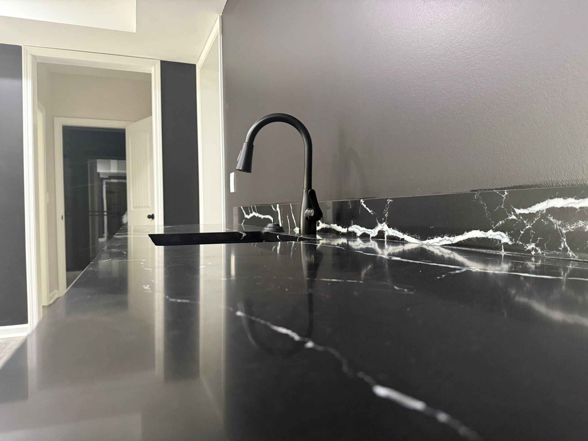 Black marble countertop with black faucet in a kitchen, hallway visible in the background.
