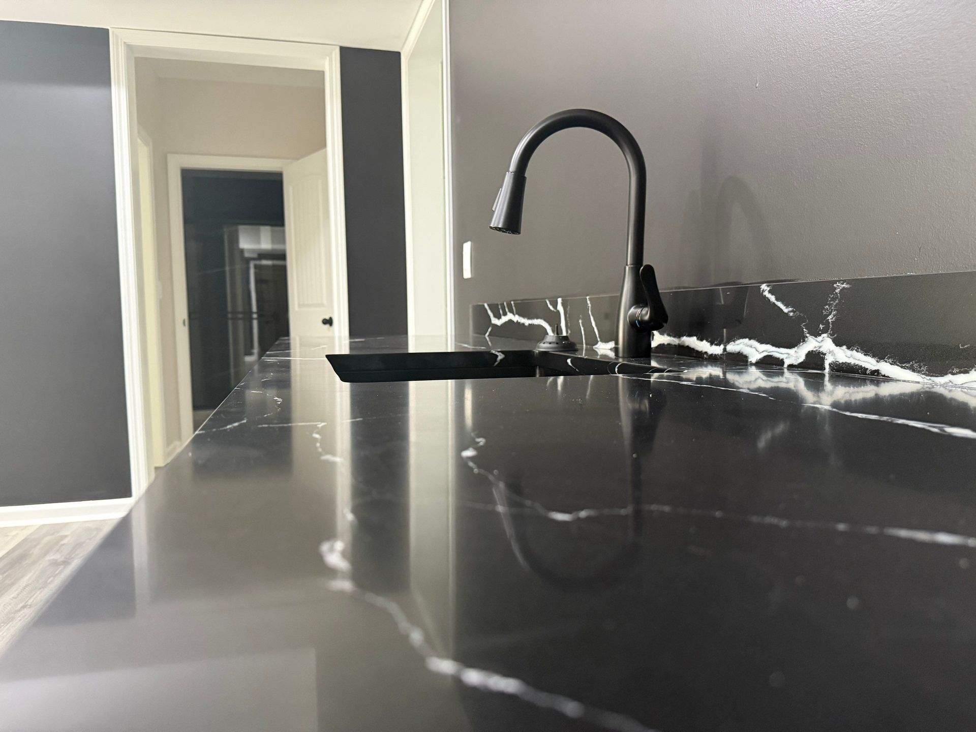 Black faucet over a sink on a black marble countertop. Hallway with dark gray walls in background.