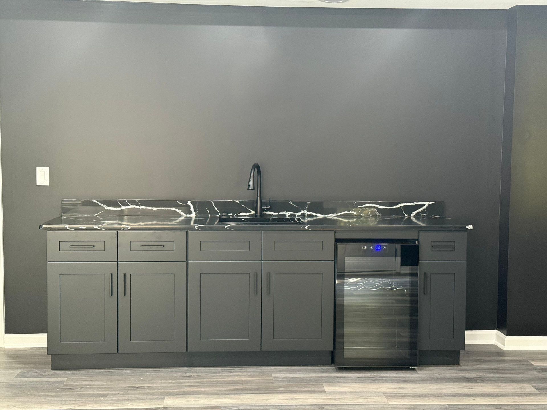 Dark gray bar with black countertop, sink, and wine fridge against a dark gray wall.