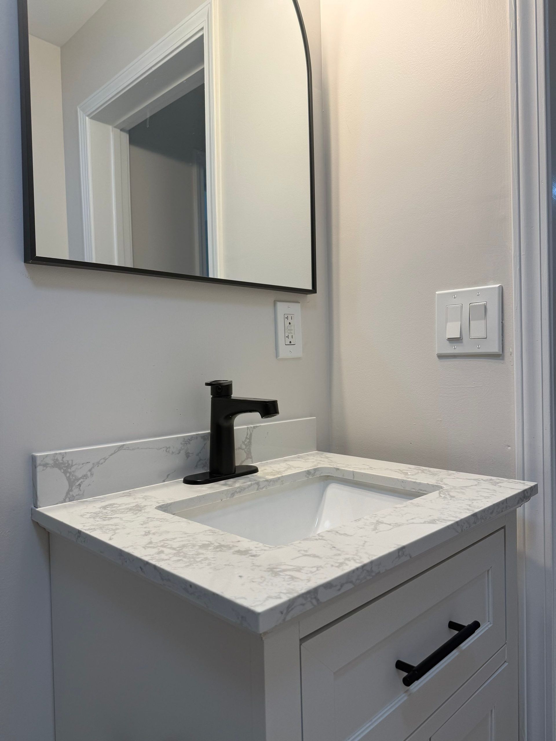 Small bathroom vanity with white countertop, black faucet, mirror, and cabinet hardware.