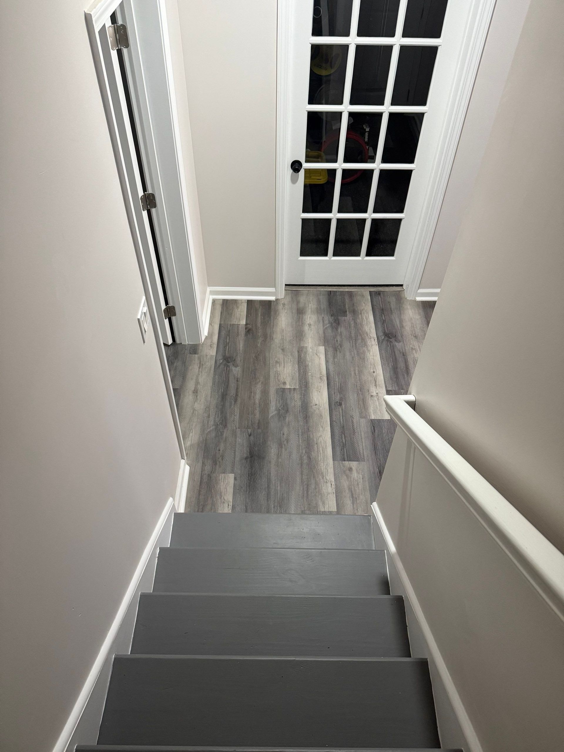 View from top of gray stairs leading to a room with gray wood-look flooring and a glass-paned door.